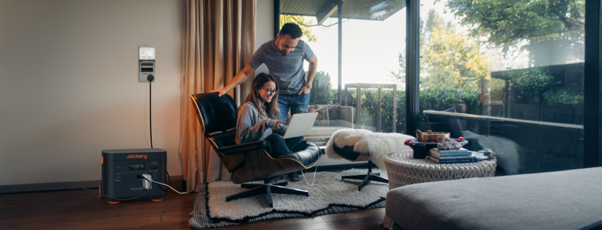 A woman sits on a chair using a laptop, with a man standing beside her in a modern living room with large windows. The Jackery Explorer 2000 Plus Portable Power Station, featuring a LiFePO4 battery, is plugged into the wall, highlighting sustainability through solar charging and lush greenery outside.