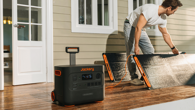 A person is setting up a portable solar panel on a wooden deck, with a Jackery Explorer 3000 Pro in the foreground featuring input and output ports. A house with a white door and windows is visible in the background.