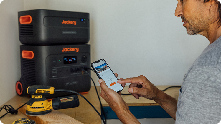 Using a phone app, someone controls two Jackery Explorer 2000 Plus power stations with LiFePO4 batteries, stacked on a wooden shelf. Below them, a DeWalt sander is ready for use, illustrating the remote management and efficiency of portable power sources.