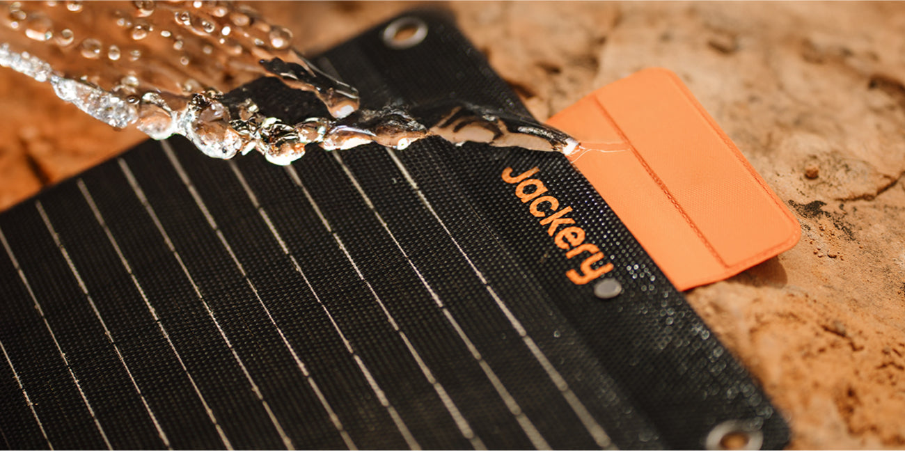 A close-up of the Jackery Solar Generator 300 Plus + SolarSaga 40W shows a stream of water flowing over its textured surface, highlighting its water resistance. The panel has black grid lines, an orange tab with mounting eyelets, and multiple charging ports for versatility.