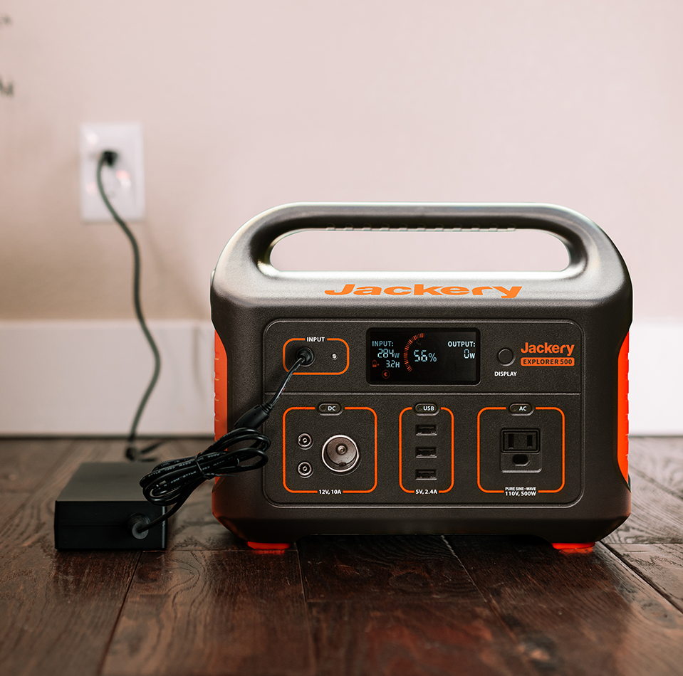 The Jackery Explorer 500, black with orange accents, rests on a hardwood floor near an outlet. It offers a high battery capacity with USB and AC ports. The small display shows 56% battery level while connected to a charger.