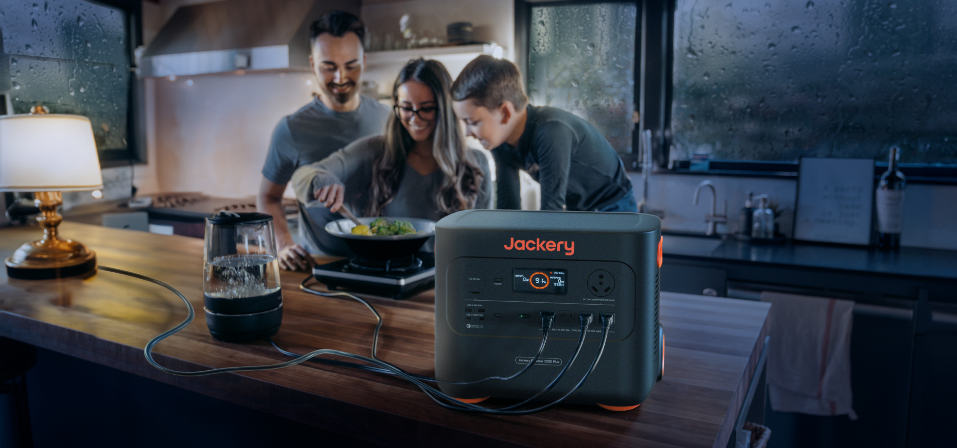 In a cozy kitchen, three people cook while the Jackery Explorer 2000 Plus Portable Power Station powers a blender and another device on the counter. Outside, silent rain falls as warm indoor lighting enhances the comforting atmosphere.