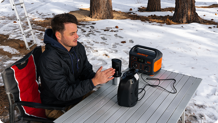 A man sits outdoors at a snow-covered folding table, using the Jackery Explorer 500 Portable Power Station to run a small electric device. The table also holds a travel mug and lantern, while trees and a ladder stand in the background.