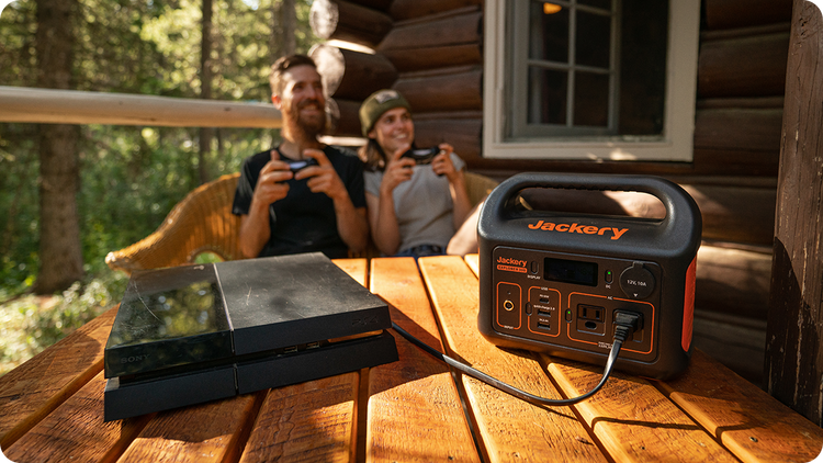 A Jackery Explorer 300 Plus Portable Power Station rests on a wooden table outside a log cabin, powering a gaming console. In the background, two people lounge in wicker chairs with game controllers, as sunlight filters through the trees and dances across the window—a perfect blend of nature and technology.