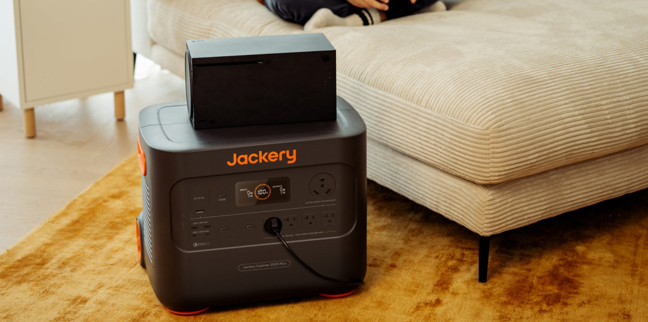 A Jackery Explorer 2000 Plus Portable Power Station is beside a beige sofa, topped with a black device, on warm light wood floors. A person's legs rest on the sofa, showcasing the convenience of ultra-fast solar charging for relaxed living.