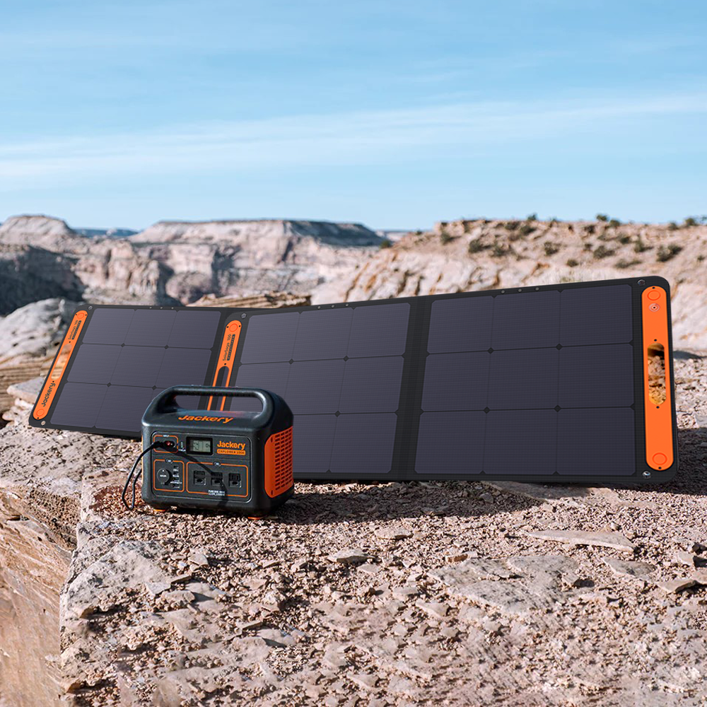 A Jackery Explorer 500 power station and solar panel sit on a rocky desert surface. The panel, open and tilted for optimal sunlight, boosts battery capacity against the rugged cliffs and blue sky.
