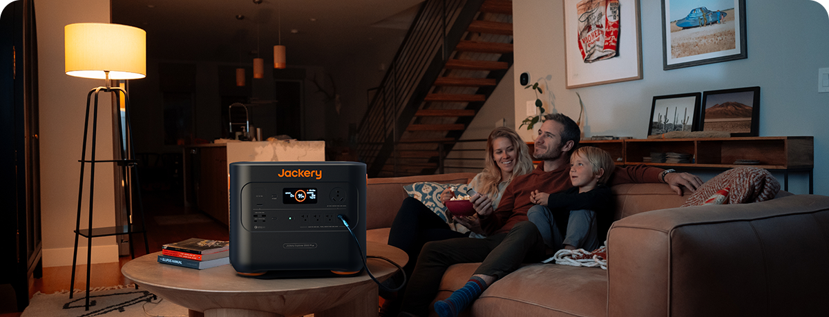 A family of three relaxes on a cozy couch in a warmly lit living room, watching something. The Jackery Explorer 2000 Plus Portable Power Station sits on the small round table in front of them, its modern design accentuating the decor with a staircase beautifully visible in the background.