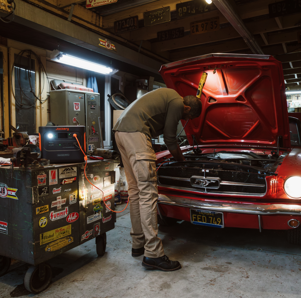 In a garage adorned with old license plates and overhead lighting, a person works on the engine of a red vintage car beside an open hood. Nearby, a sticker-covered toolbox and the Jackery Explorer 1000 Plus Portable Power Station provide expandable capacity for all power needs by the workbench.