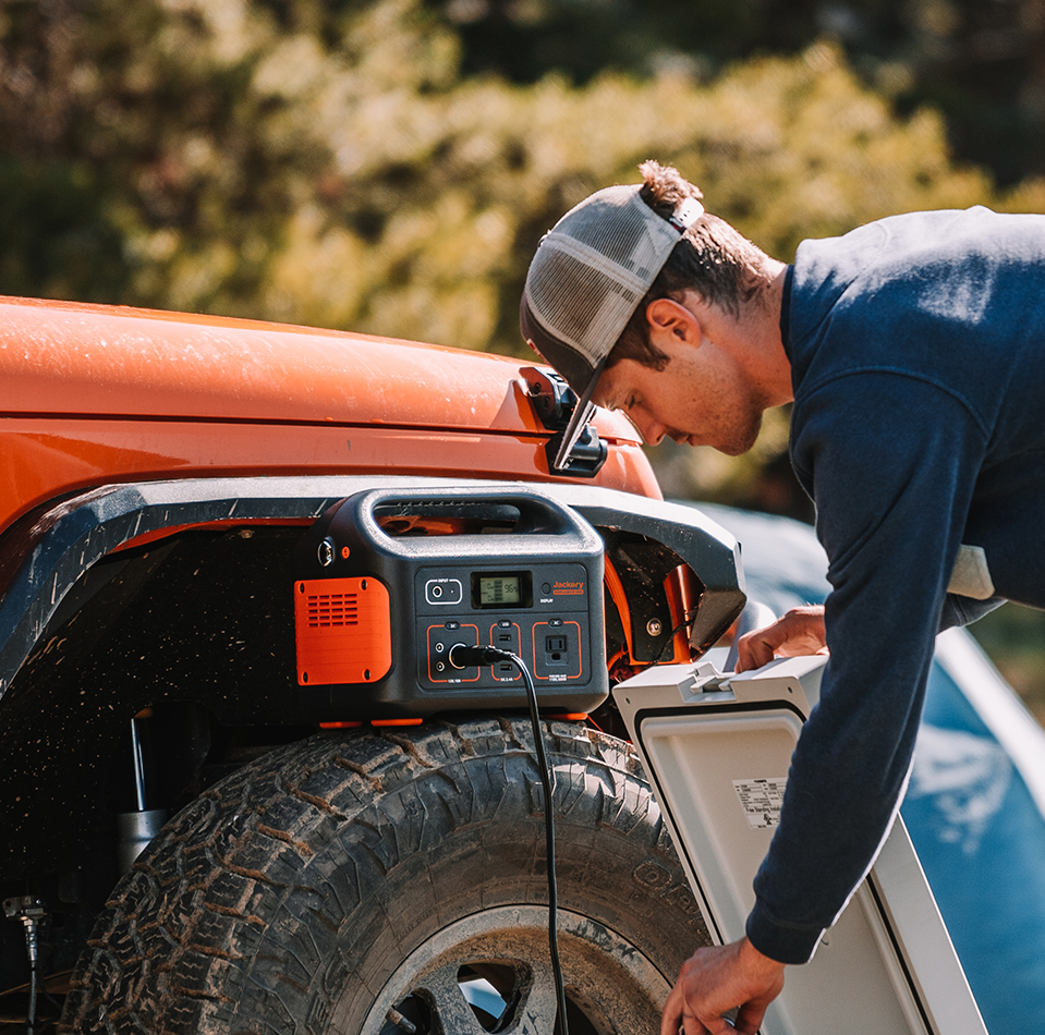 A person in a baseball cap uses the Jackery Explorer 500 Portable Power Station, attached to an orange SUV outdoors. Positioned above a large tire, the power station by Jackery blends with a blurred green backdrop.