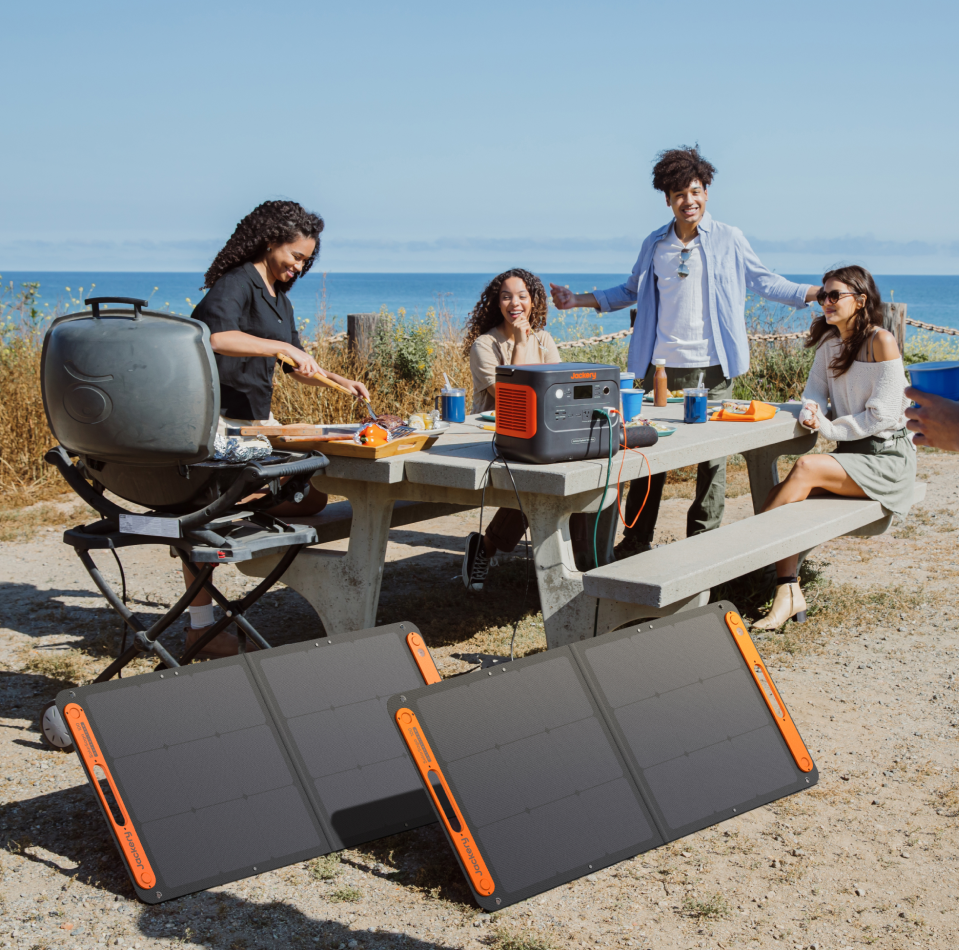 Four people enjoy a sunny day by the ocean at a picnic table, using a Jackery Explorer 1000 Plus Portable Power Station connected to solar panels. One person stands by a grill in this outdoor setting, with clear skies and a scenic sea view., 2000wh power station, cpap portable power station