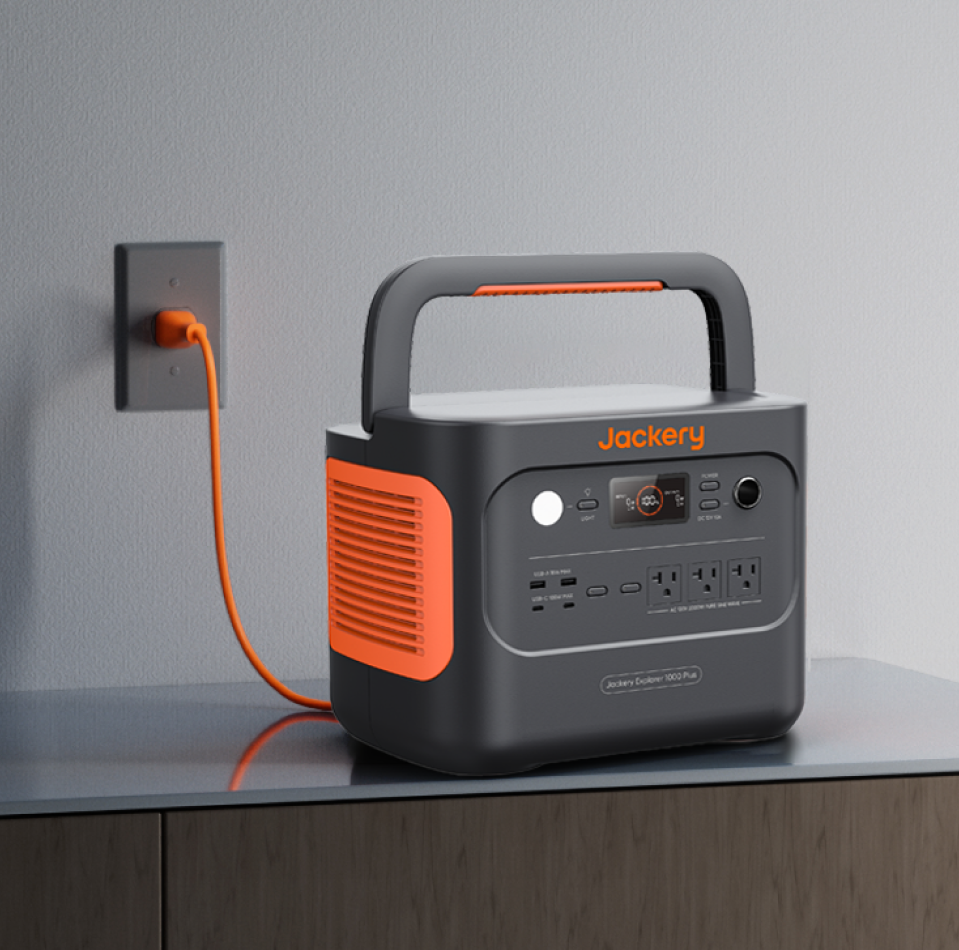 The Jackery Explorer 1000 Plus Portable Power Station rests on a wooden surface, connected to a wall outlet with an orange cord. It includes AC outlets, USB ports, a display screen, and offers expandable capacity for various needs. The device is gray with orange accents.